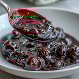 Silky smooth Black Currant Sauce drizzled generously over a slice of creamy vanilla cheesecake on a dessert plate.