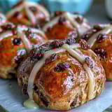 Golden hot cross buns topped with orange glaze, warm spices, and sweet dried fruit in a cozy kitchen scene.  